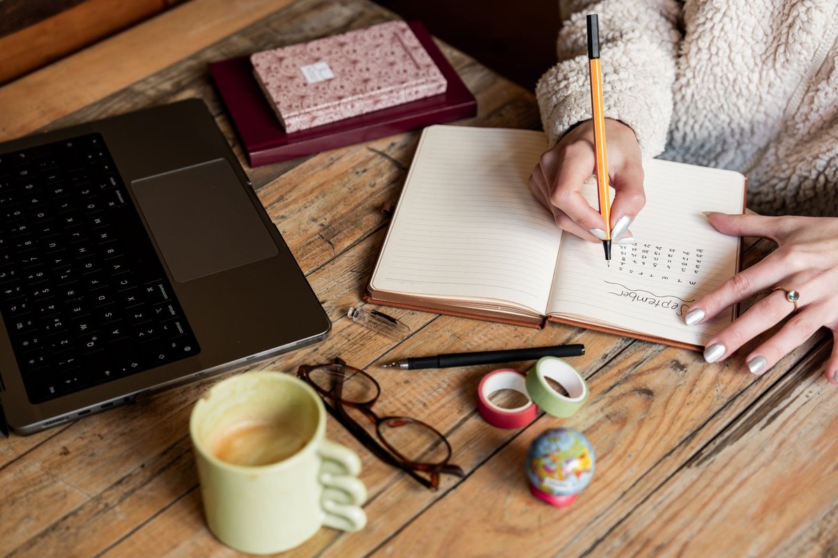 A woman writing in a notebook beside a laptop and coffee