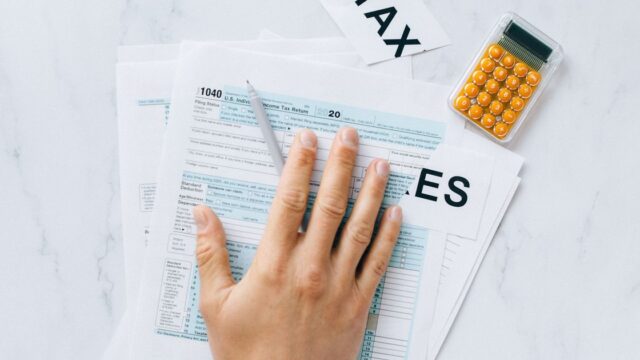 A hand resting on a US tax form with a calculator on the desk