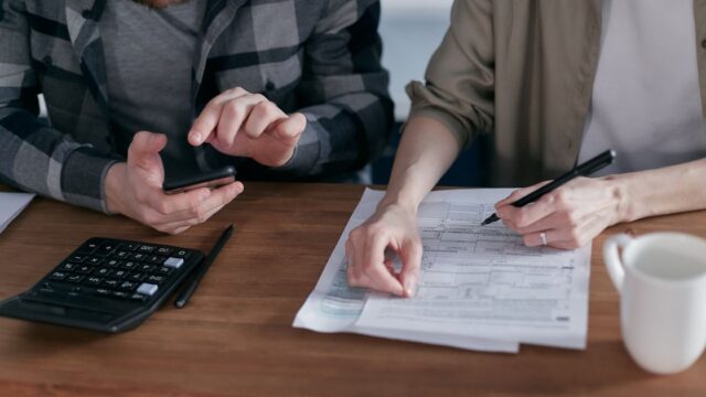 Two people filling in tax forms together at a wooden desk with a calculator