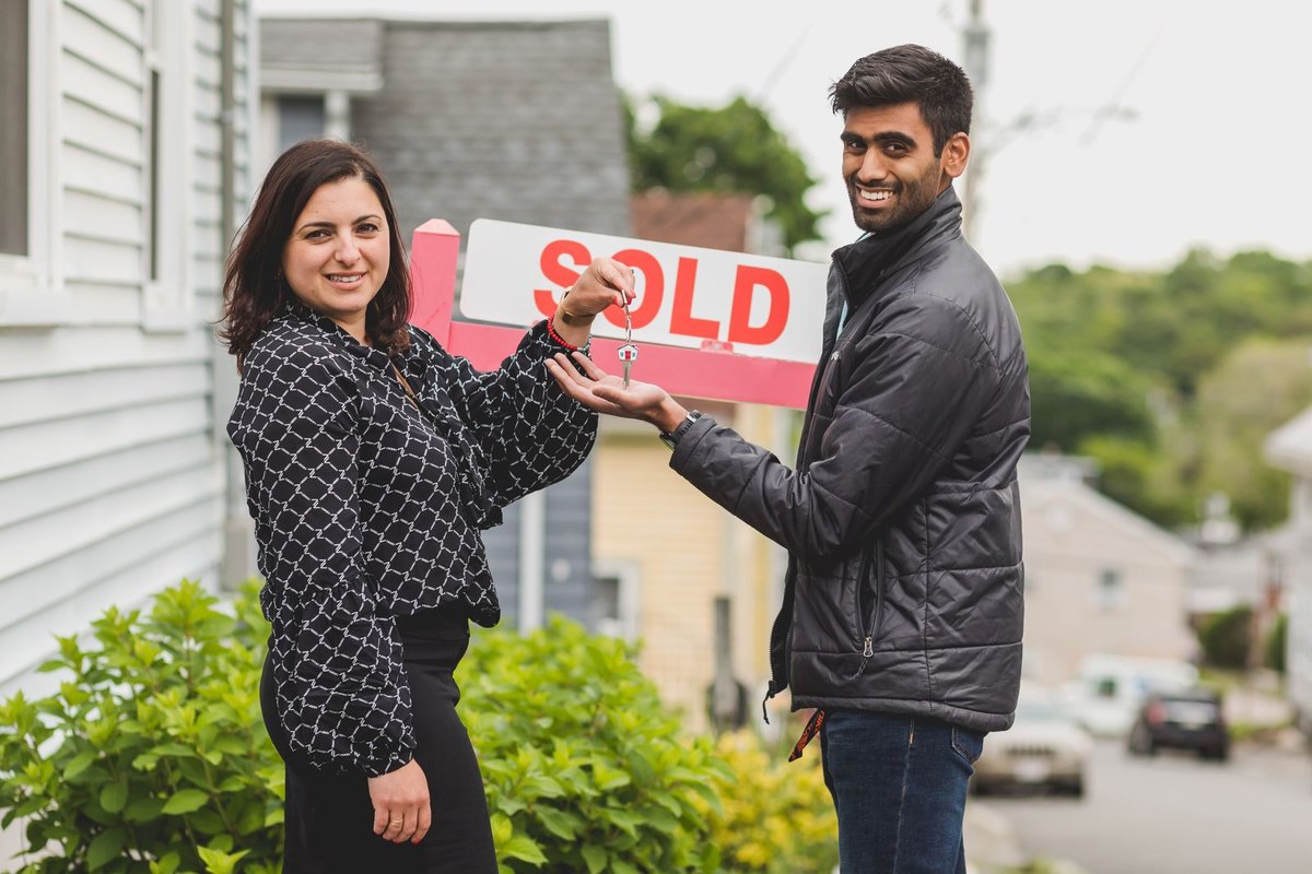 A real estate agent handing a house key to a couple standing in front of a sold sign