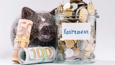 A glass jar of coins labelled Retirement beside a piggy bank and rolls of banknotes