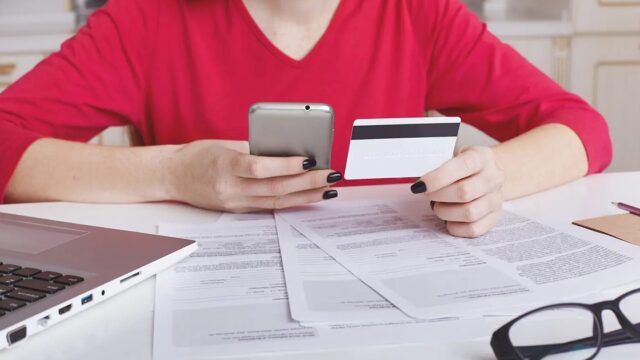 A woman at a desk holding a phone and a bank card above loose paperwork