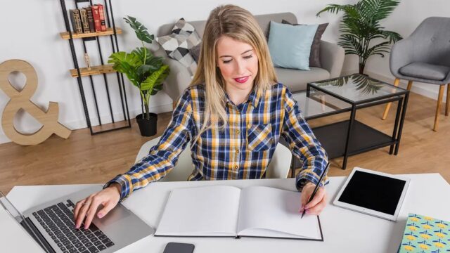 A woman at a table with a laptop and an open notebook in a bright living room