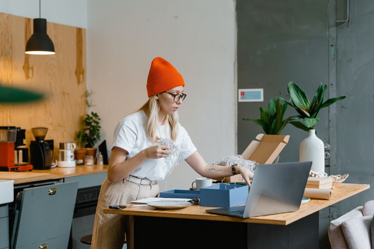 A woman in a red beanie packing an online order at a table with a laptop