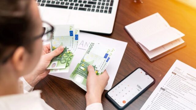 A woman counting euro notes at a wooden desk with a laptop and paperwork