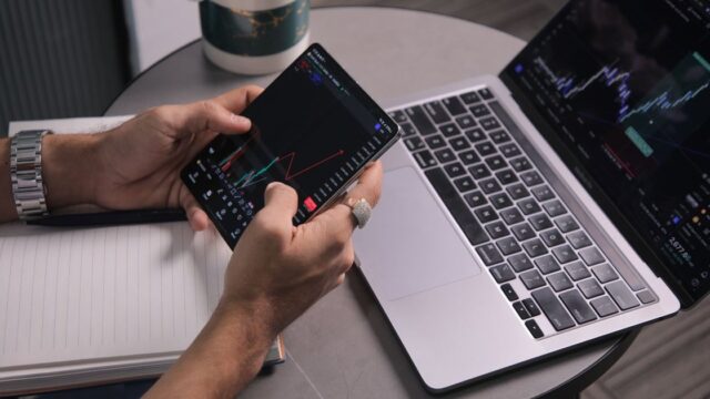 A man looking at market charts on a laptop and a trading app on his phone