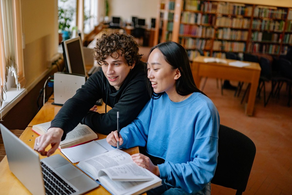 Two students studying together in a library with a laptop and books