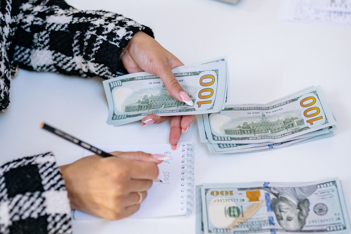 A woman counting dollar bills with a pencil in hand beside a laptop