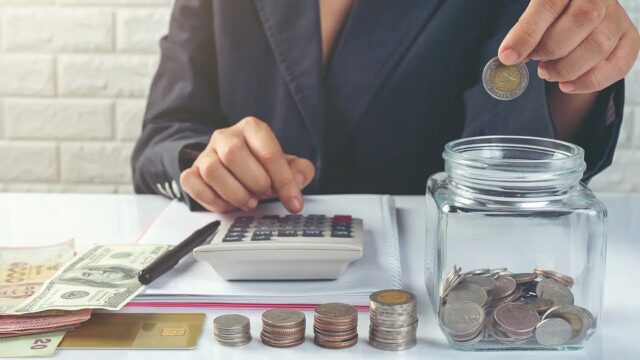 A woman dropping a coin into a jar beside a calculator and banknotes