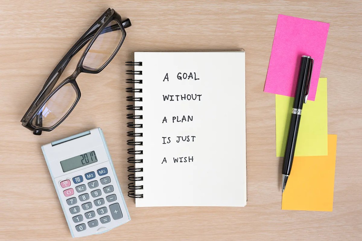 A notebook reading 'A goal without a plan is just a wish' with glasses, a calculator and sticky notes