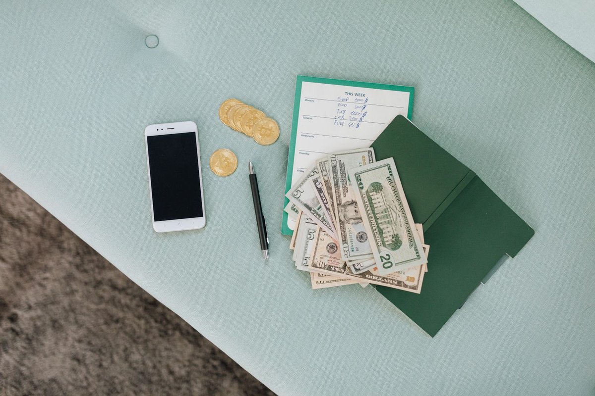 A smartphone, dollar bills, coins and a planner on a green table
