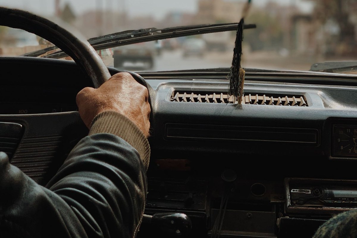 A man's hand on the steering wheel of an older car while driving through a city