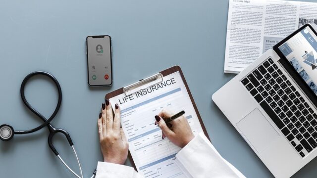 A doctor's hands filling in an insurance form next to a stethoscope, phone and laptop